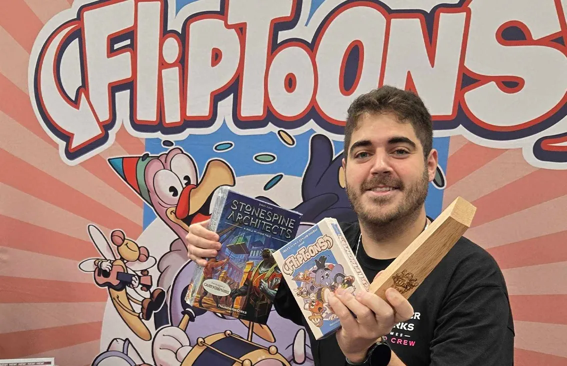 Na foto: Renato Simões (eu), segurando o troféu de Staff Pick da Gen Con 2025, junto com uma cópia do FlipToons e, representando o outro game designer responsável pelo jogo, Stonespine Architects.